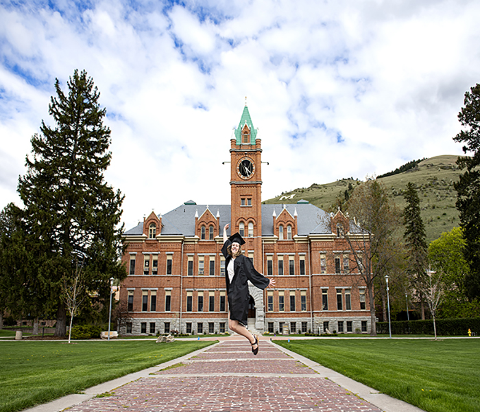 student jumping in front of main hall in a graduation robe 